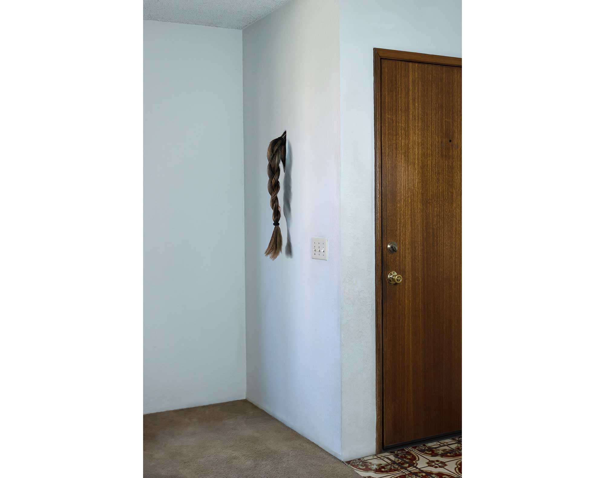 A braided ponytail emerges from the wall in the corner of a room