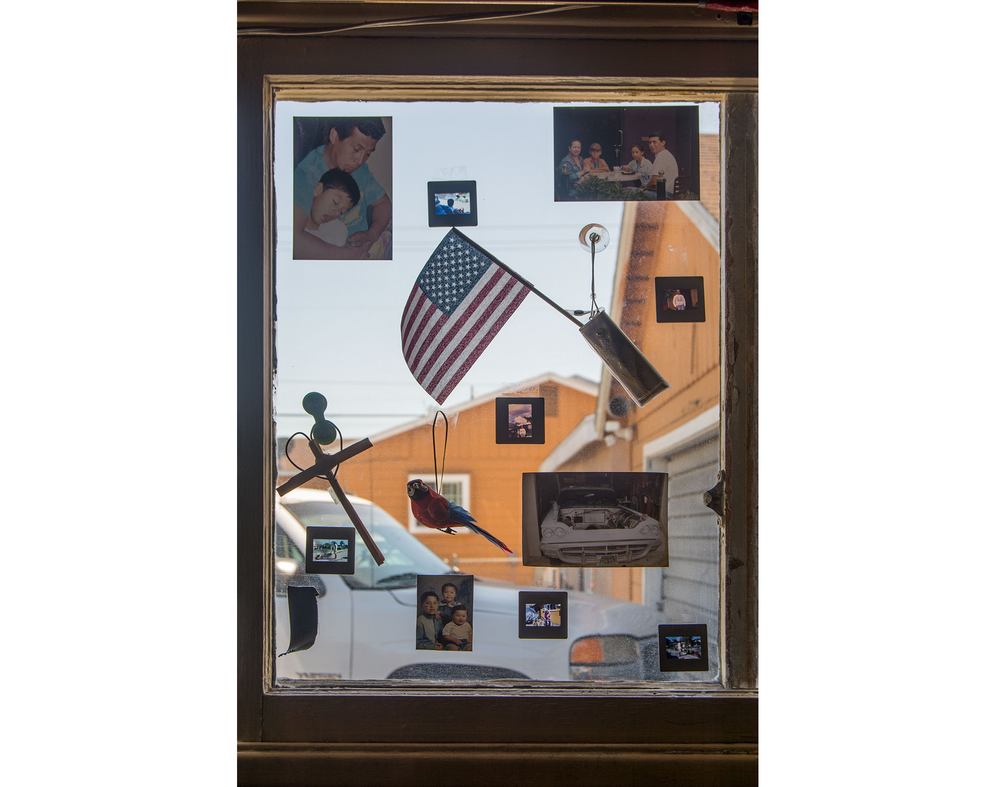 A photograph of a window that has pigmented photographs taped on the glass. 35mm slide film is also mounted on the glass along with an ornament of a parrot. There is a cross made by palm leaves mounted to the glass by magnets. There is an American flag inside a sleeve using a suction cup.