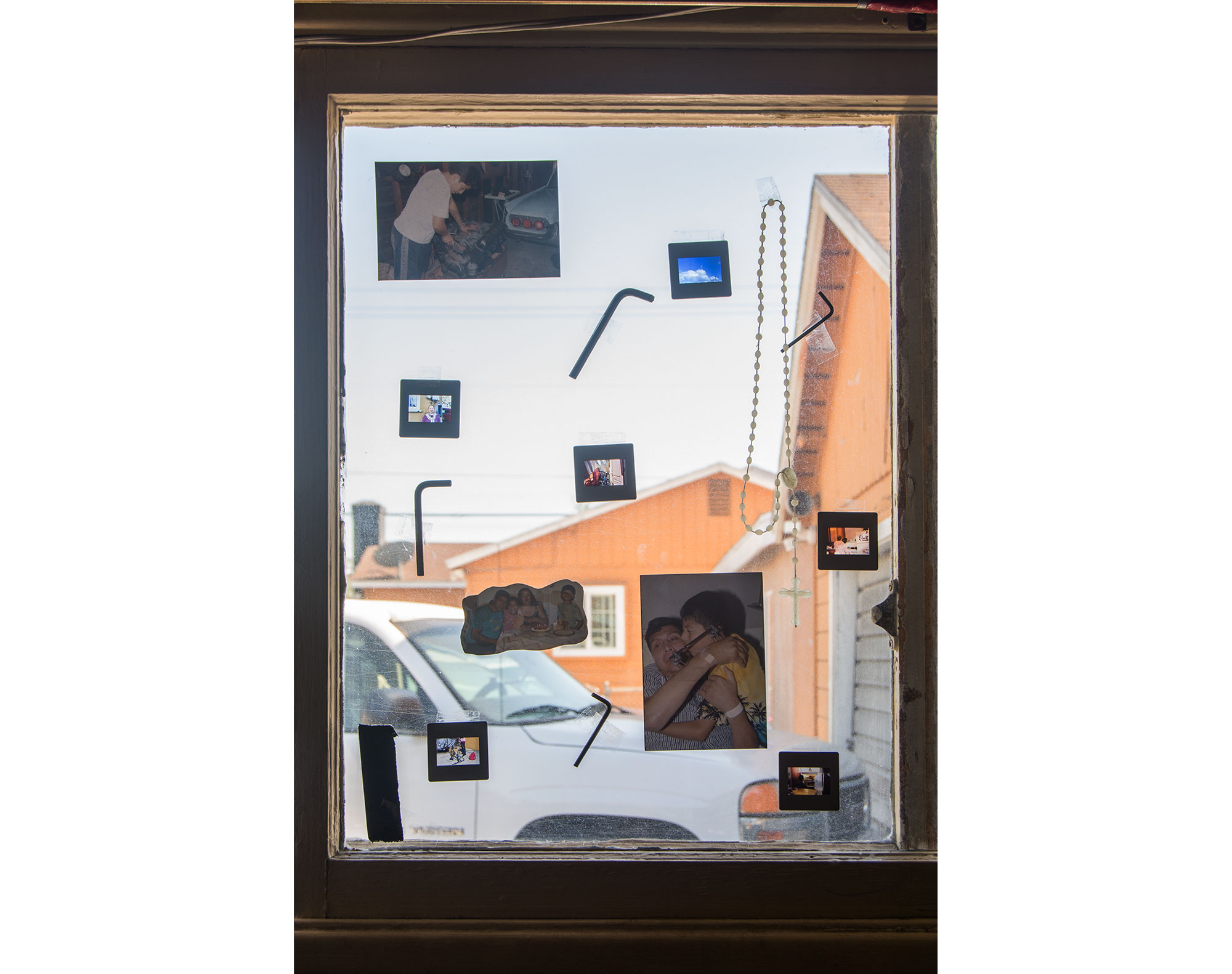 A photograph of a window that has pigmented photographs taped on the glass. 35mm slide film is also mounted on the glass with tape along with a rosary and allen wrenches taped all over the glass. One of the smaller photographs has my father at the hospital hugging me. The second photograph is my brother working on the engine of a 1960 Thunderbird.