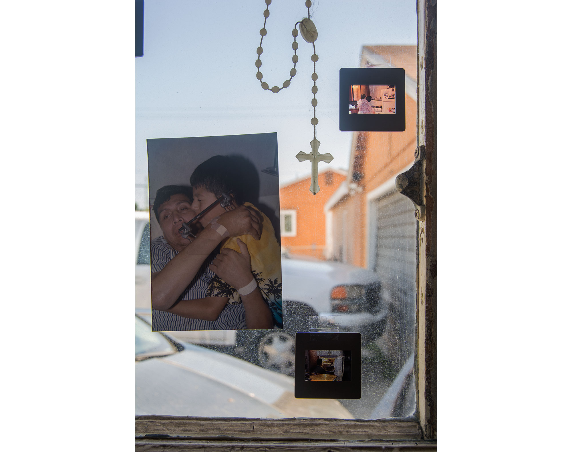 A close up shot of the first piece that focuses on the cross made of palm leaves, red parrot, 35mm slide, and part of the American flag. There is also a wallet sized photograph of my brother, sister, and I