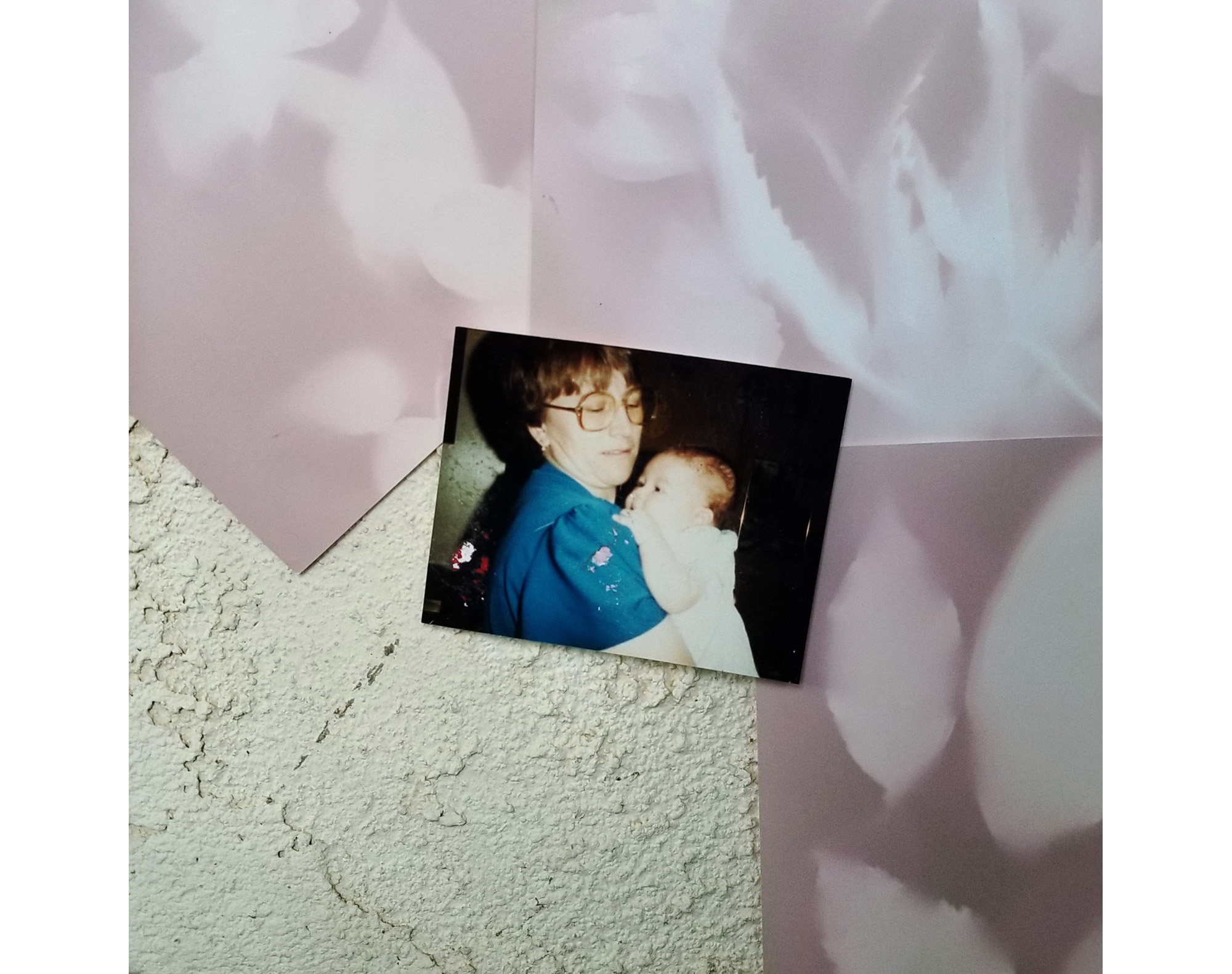 Close up of photo showing Mom holding infant placed over lumen prints. Mom in blue and infant in white dress. White and light pink images of flowers and plants are formed over pastel pink, and purple background. The textured wall in the left corner is exposed.  